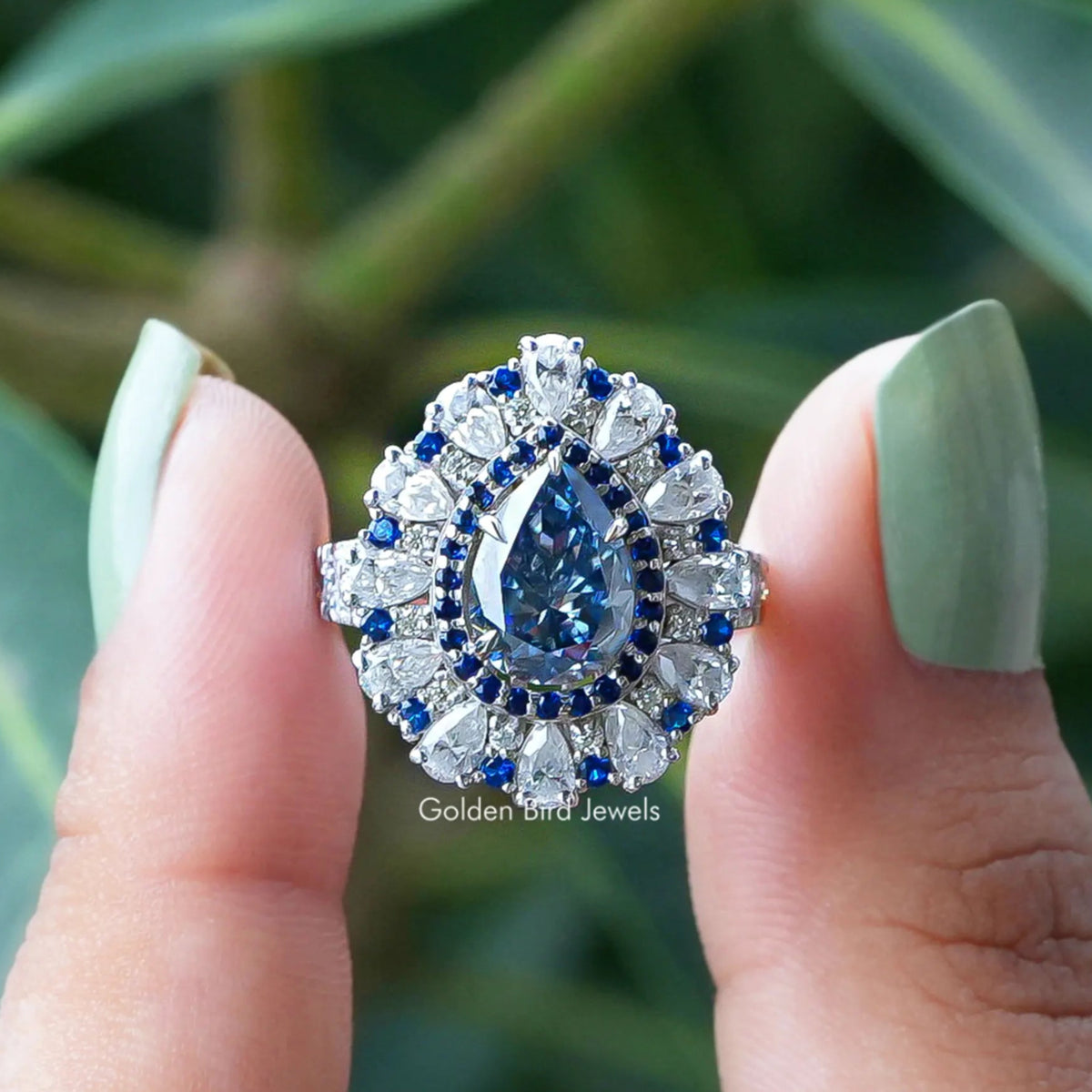 Two fingers holding the Blue Pear Cut Moissanite Halo Engagement Ring