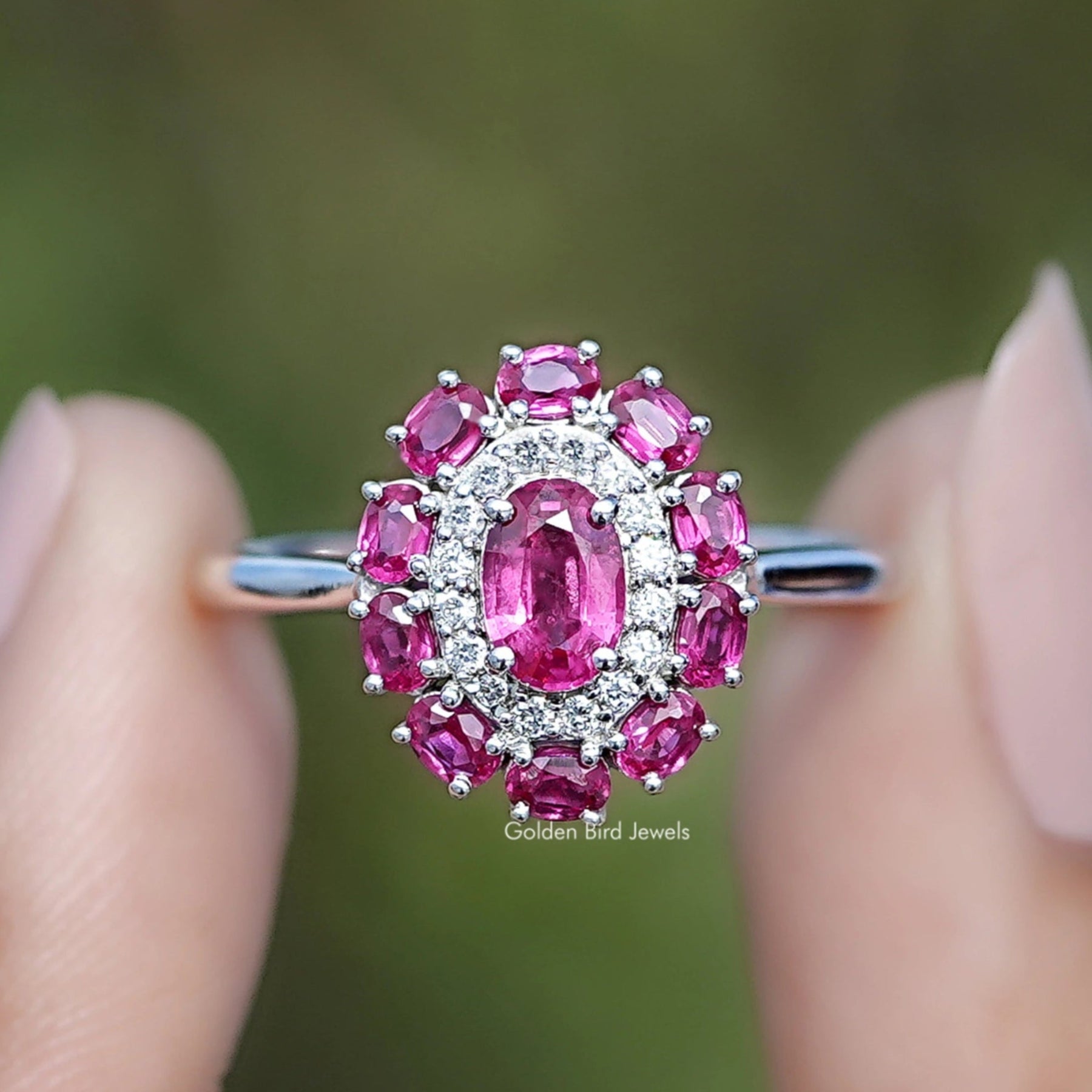 Pink gemstone ring held between two fingers with a blurred green background
