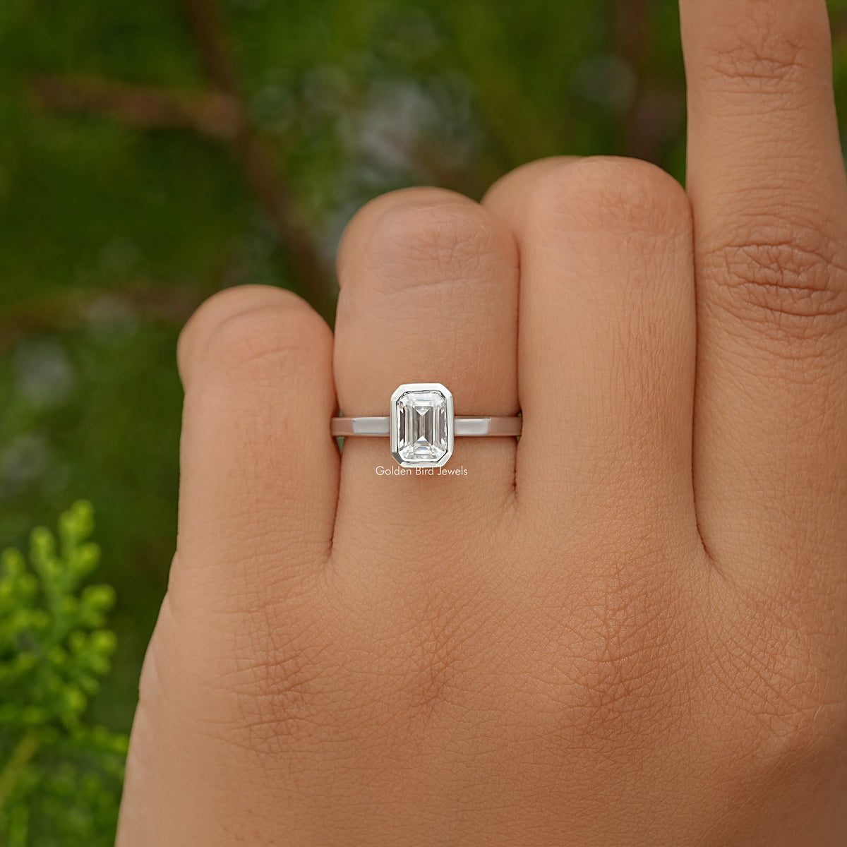 Emerald-cut moissanite bezel-set engagement ring on hand, front view of the stone