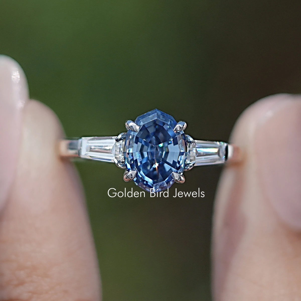 Front view of Blue Step Cut Oval Moissanite Three Stone Ring in two fingers 