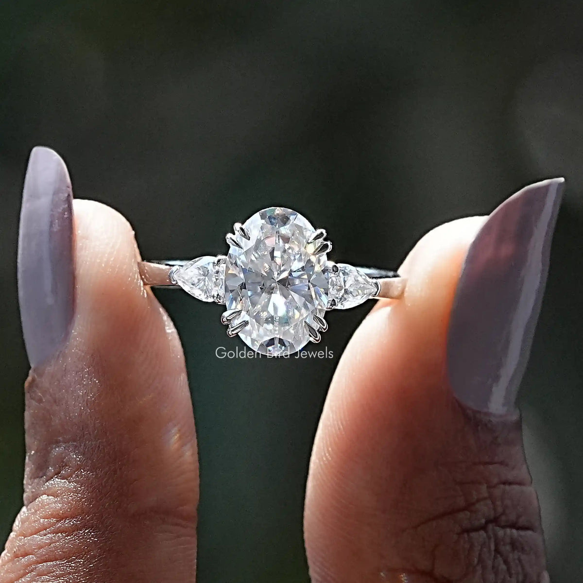 Two fingers holding the Oval And Pear Cut Moissanite 3 Stone Engagement Ring
