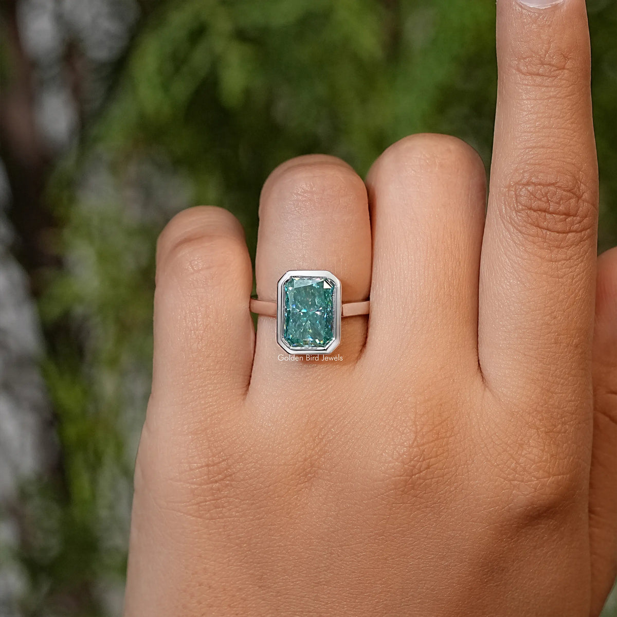 Teal emerald-cut moissanite solitaire ring on hand, bezel setting visible