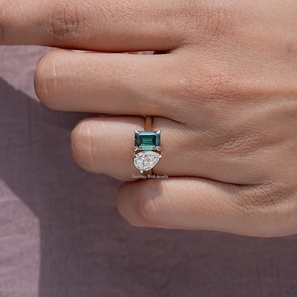 Close-up two-stone ring: emerald-cut green stone and round white stone, prongs detailed